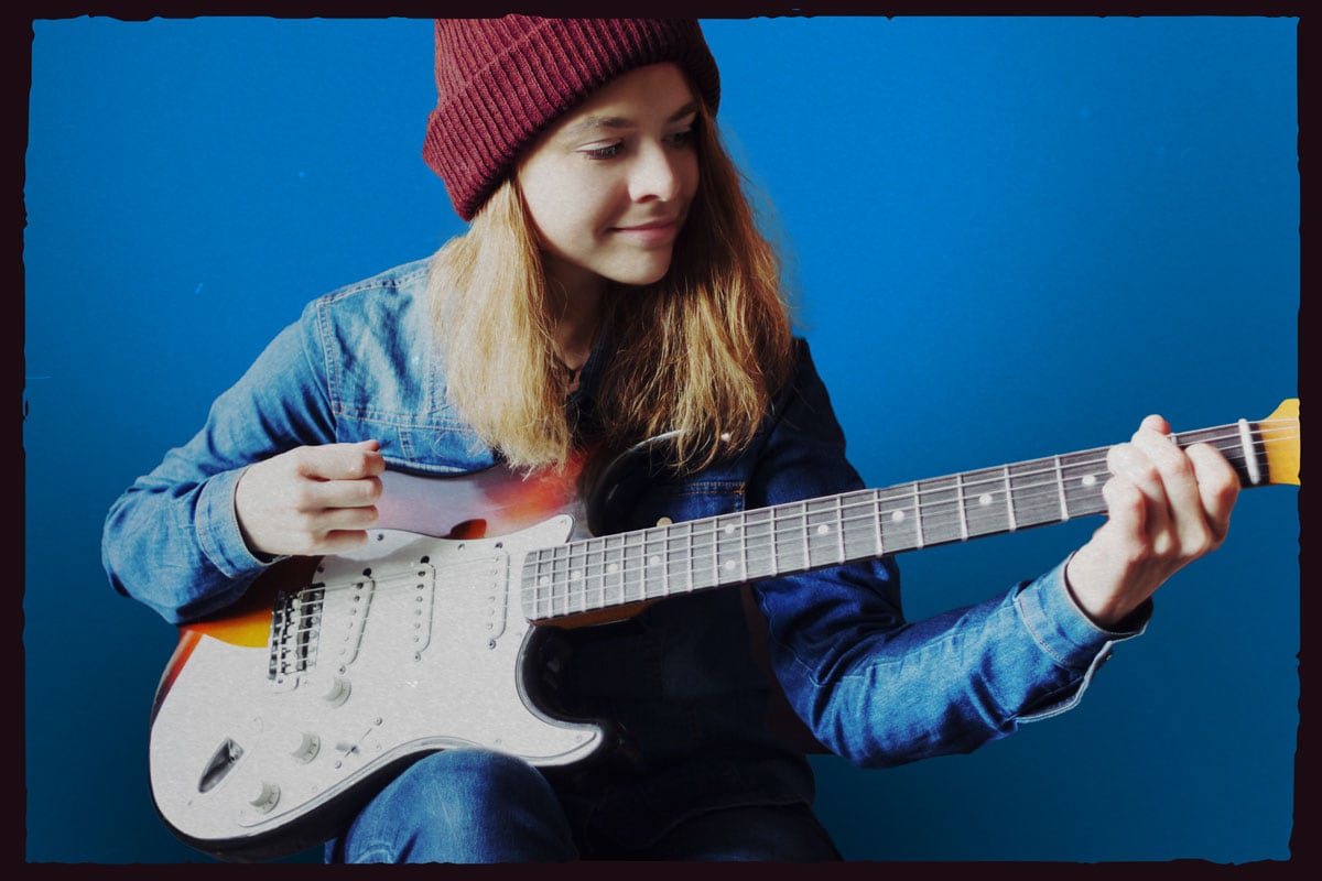 Female teen wearing a maroon knit cap and jean shirt strumming an electric Stratocaster guitar.