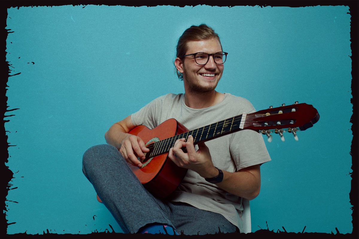 Twenty-something man with tortoise shell glasses and his hair tied back sitting with an acoustic guitar.