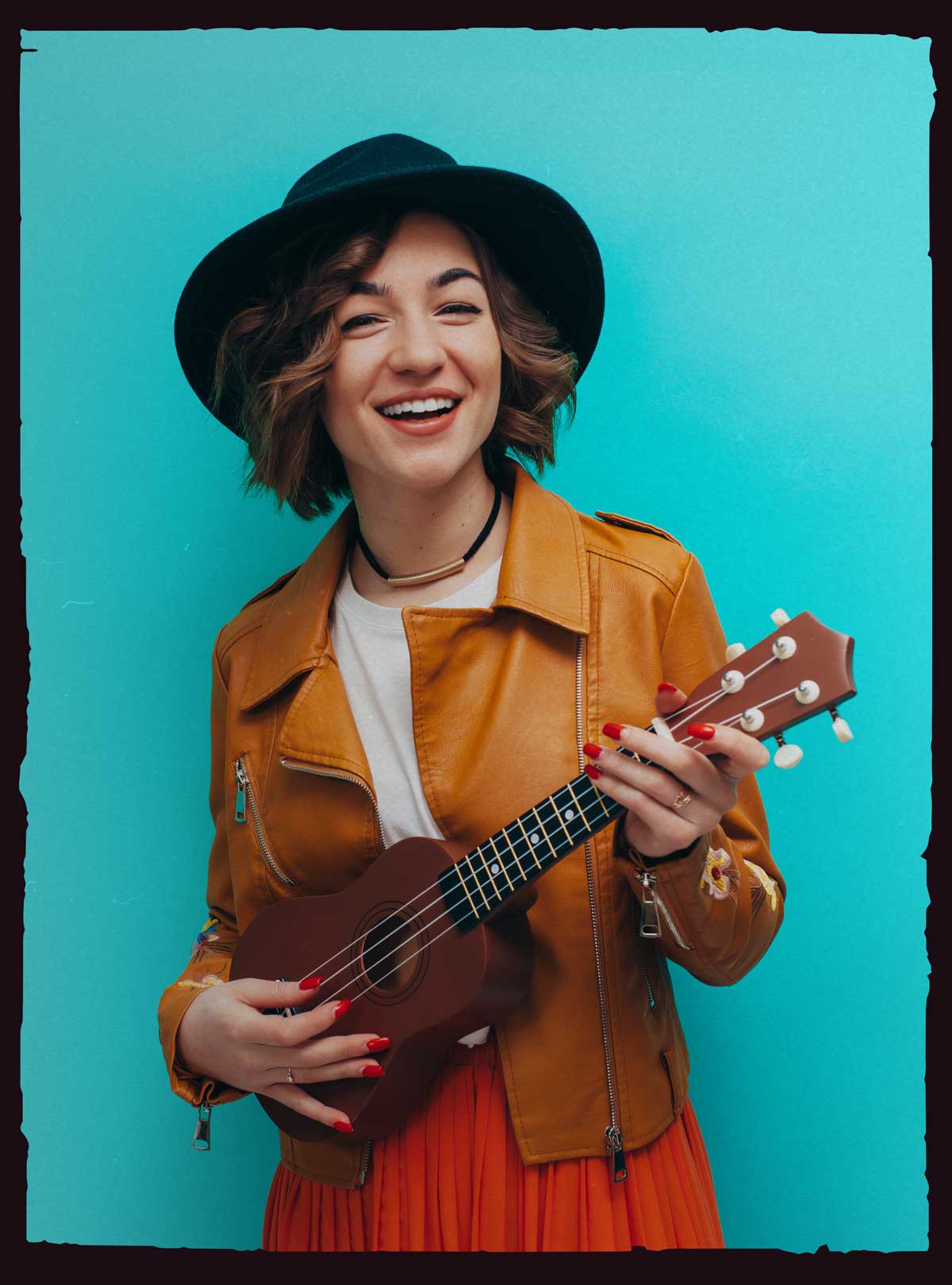 Twenty-something female wearing a tan leather motorcycle jacket and blue felt fedora holding a ukulele