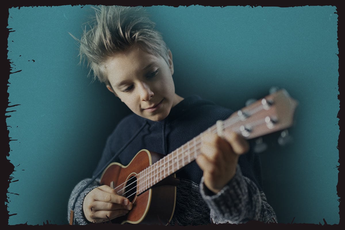 A tween boy with navy hoodie concentrating on playing an ukulele.