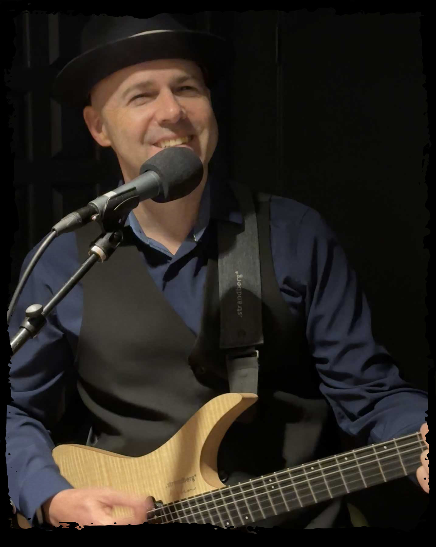 Barrett Simms wearing a navy shirt and black vest smiling at a microphone with his maple electric Strandberg guitar.